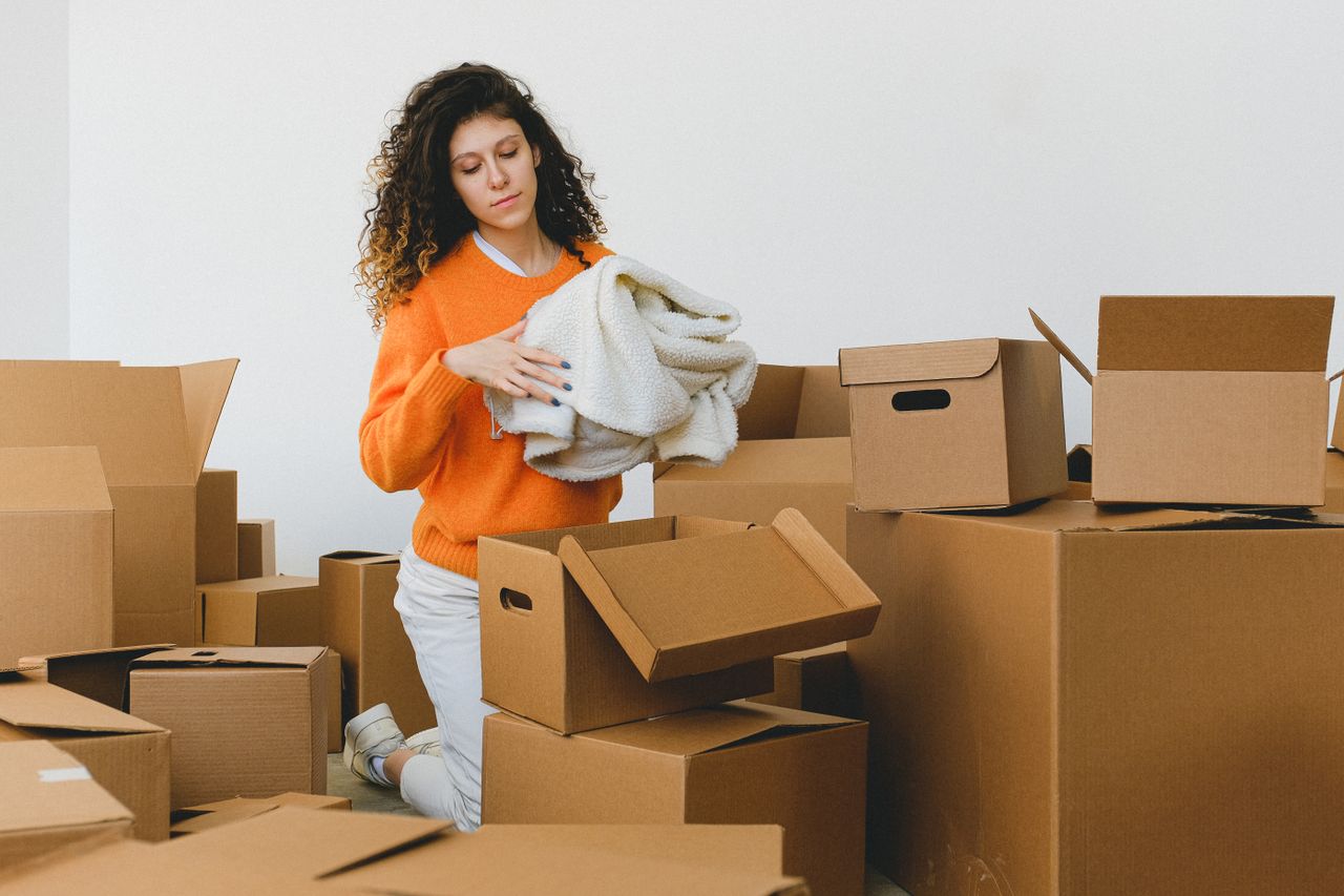 about-01 Concentrated young lady with curly hair standing on knees on floor and packing clothes into carton boxes before moving into new flat