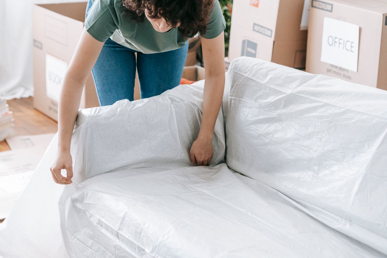 services-09 A woman carefully wraps a sofa in protective material while packing boxes in a bright room.