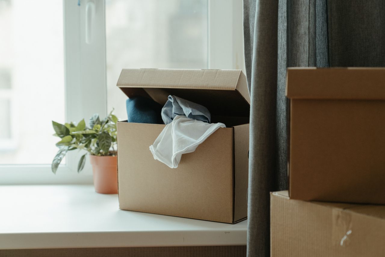 hero-01 Open cardboard box with clothes by window, symbolizing moving or relocation.